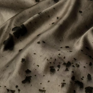 Peut inclure: Gros plan sur un tissu beige parsemé de taches irrégulières brun foncé. Le tissu est drapé, créant des plis et des ombres douces. La texture semble lisse avec un léger éclat, suggérant un matériau délicat.