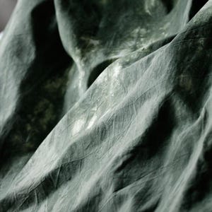 Puede incluir: Primer plano de una tela drapeada en tonos verdes y grises. La tela tiene una apariencia texturizada con pliegues visibles y sutiles variaciones de color, creando una sensaci&oacute;n de profundidad y movimiento. La est&eacute;tica general es suave y org&aacute;nica.