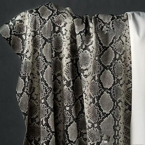 Pode incluir: Um tecido drapeado com um padrão de pele de cobra em tons de cinza, preto e branco. O tecido texturizado tem dobras e sombras visíveis, sugerindo um material pesado. Um tecido branco liso é visível no lado direito.