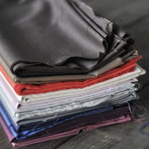 May include: A stack of various colored fabric swatches. The fabrics include shades of gray, red, white, blue, and purple. The edges of the fabric are unfinished, giving a raw, textured appearance. The fabrics are stacked on top of each other.