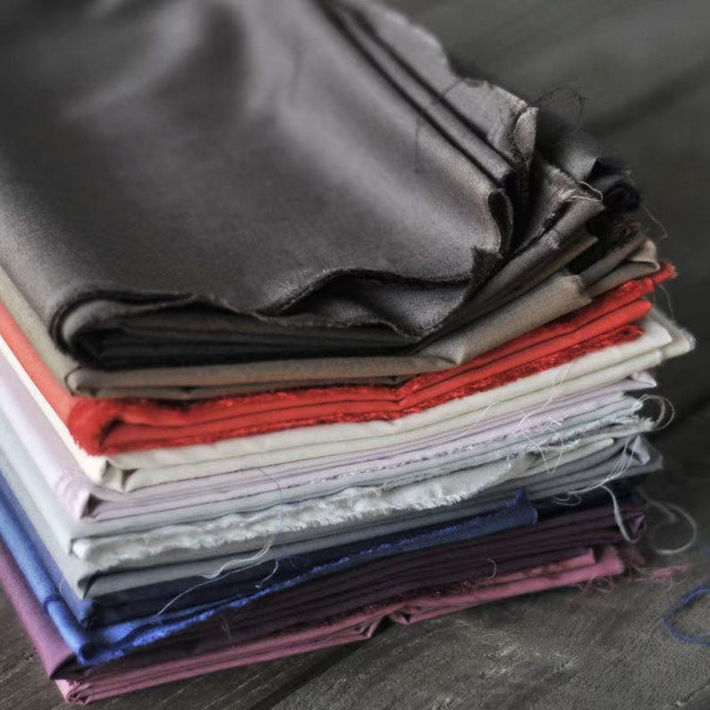 May include: A stack of various colored fabric swatches. The fabrics include shades of gray, red, white, blue, and purple. The edges of the fabric are unfinished, giving a raw, textured appearance. The fabrics are stacked on top of each other.