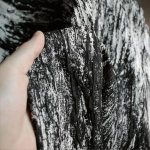 Puede incluir: Primer plano de una tela texturizada en blanco y negro. La tela tiene un aspecto arrugado con un patrón que se asemeja a pinceladas. El material se sostiene, revelando su textura y diseño.