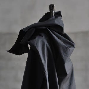 Könnte beinhalten: Ein dunkelmarineblauer Stoff, der über einem Holzständer drapiert ist. Der Stoff ist zerknittert und gefaltet, wodurch Schatten und Textur entstehen. Der Ständer hat eine dunkle, dekorative Oberseite. Der Hintergrund ist ein neutrales Grau.