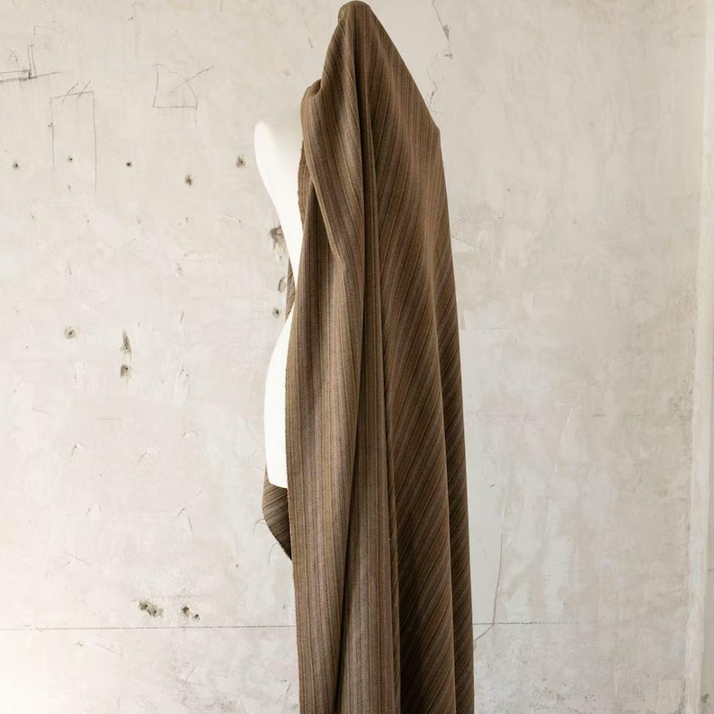 May include: Brown fabric with vertical stripes draped over a white mannequin. The fabric has a textured appearance and a warm, earthy color palette. The stripes vary in width and shade, creating visual interest.