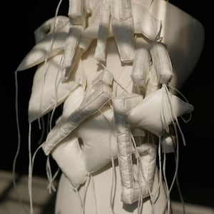 Puede incluir: Prenda abstracta de color blanco roto con un diseño complejo. La pieza presenta numerosas formas acolchadas, rectangulares y triangulares, conectadas por finos cordones blancos. La textura general parece suave y acolchada, con un enfoque en las formas geométricas y una estética minimalista.