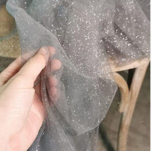 Könnte beinhalten: Nahaufnahme eines grauen Tüllstoffs mit silbernen Glitzerpartikeln. Der Stoff wird hochgehalten, wodurch seine Textur und die Art und Weise, wie er das Licht einfängt, sichtbar wird. Der Tüll ist über einen Holzstuhl drapiert.