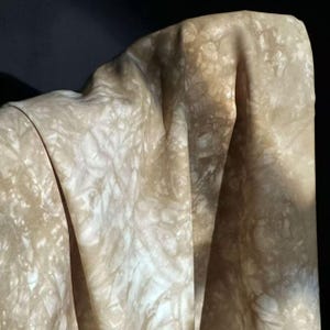Puede incluir: Una tela drapeada con un estampado tie-dye en tonos beige y blanco roto. La tela es suave y fluida, con pliegues y sombras visibles que crean profundidad y textura. El efecto general es orgánico y natural.
