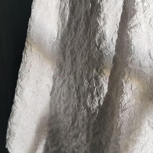 May include: Close-up of white fabric with a textured, crinkled surface. The fabric appears to be draped, with folds and shadows creating depth. The material has a subtle sheen, reflecting light and adding visual interest. The overall effect is one of delicate texture and subtle elegance.