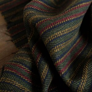 May include: Close-up of a green fabric with a herringbone pattern. The fabric features stripes in red, yellow, and blue. The edges of the fabric are frayed, suggesting a scarf or textile.
