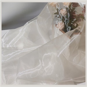 Può includere: Un tessuto bianco sporco e trasparente con una sottile lucentezza, drappeggiato e piegato. La trama del tessuto è visibile, catturando la luce e creando riflessi. Un bouquet di rose rosa e crema è parzialmente visibile sullo sfondo.