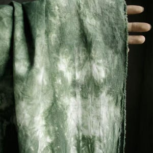 Puede incluir: Primer plano de una tela verde y blanca te&ntilde;ida con la t&eacute;cnica tie-dye. La tela tiene un aspecto texturizado con una mezcla de tonos verdes claros y oscuros. Los bordes de la tela son visibles, mostrando un borde crudo e inacabado. La tela est&aacute; drapeada, creando pliegues y sombras.