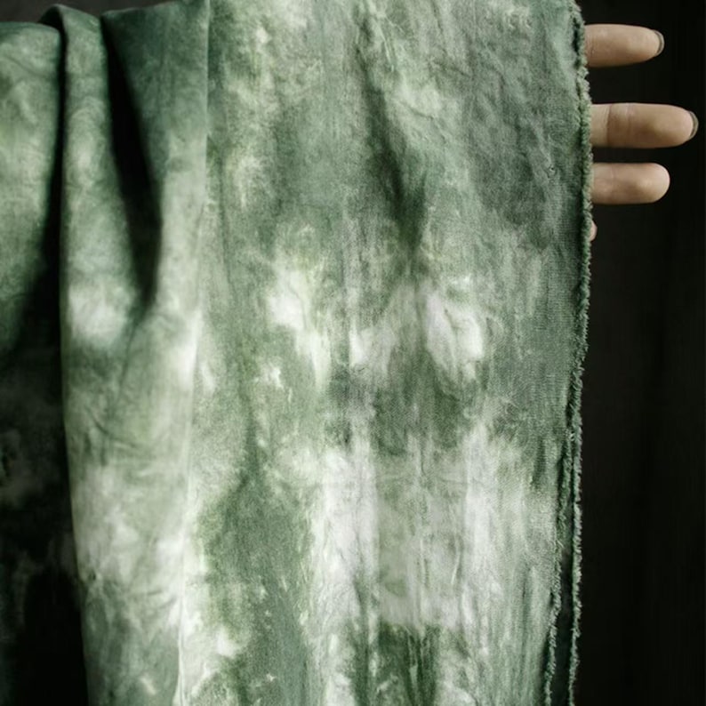 Puede incluir: Primer plano de una tela verde y blanca te&ntilde;ida con la t&eacute;cnica tie-dye. La tela tiene un aspecto texturizado con una mezcla de tonos verdes claros y oscuros. Los bordes de la tela son visibles, mostrando un borde crudo e inacabado. La tela est&aacute; drapeada, creando pliegues y sombras.
