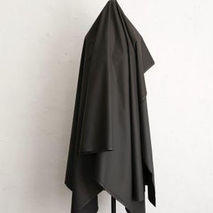 May include: A large piece of black fabric draped over a black stand against a white wall. The fabric is smooth and appears to be a solid color, with folds and shadows adding depth. The fabric is likely used for sewing or crafting.