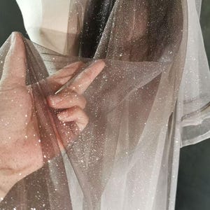 Könnte beinhalten: Nahaufnahme eines schimmernden Stoffes mit Farbverlauf, der von Dunkelbraun zu Hellbeige übergeht. Das transparente Material ist mit funkelndem Glitzer bedeckt, was einen glamourösen und ätherischen Look erzeugt. Der Stoff wird hochgehalten, um seine zarte Textur und seinen Fall zu zeigen.