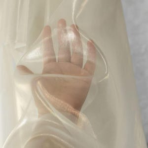 May include: A close-up of a sheer, ivory-colored fabric. The fabric is held up, creating soft folds and highlights. The material appears lightweight and translucent, with a subtle sheen.
