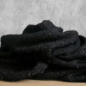 May include: Close-up of a black, textured fabric. The material appears to be thick and woven, with a slightly rough surface. The fabric is folded and layered, creating shadows and depth. The overall appearance suggests a warm, cozy textile.