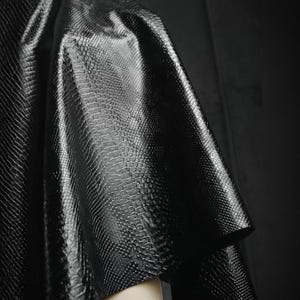 Może przedstawiać: Zbliżenie czarnej, teksturowanej tkaniny ze wzorem sk&oacute;ry węża. Materiał ma błyszczący połysk i jest udrapowany, ukazując fałdy i cienie. Tkanina wydaje się być częścią odzieży, prawdopodobnie topu lub kurtki.