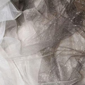 May include: Close-up of shimmering fabric in shades of white, gray, and black. The material is layered and textured, with a glittery, sparkling effect. The fabric appears soft and flowing, suitable for apparel or decorative projects.