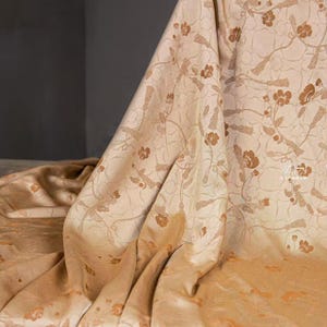 Könnte beinhalten: Beigefarbener Stoff mit einem floralen Muster in Brauntönen. Der Stoff ist drapiert und zeigt Falten und Textur. Das florale Design umfasst stilisierte Blumen und Zweige, die ein zartes und elegantes Aussehen erzeugen. Der Stoff scheint leicht bis mittelschwer zu sein.