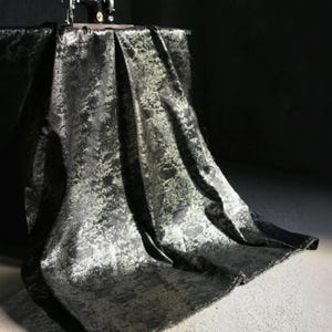 Puede incluir: Tela negra con un patr&oacute;n texturizado y moteado, drapeada sobre una mesa. La tela tiene un brillo met&aacute;lico, con reflejos m&aacute;s claros y plateados sobre el fondo oscuro. Una m&aacute;quina de coser vintage es visible sobre la mesa.