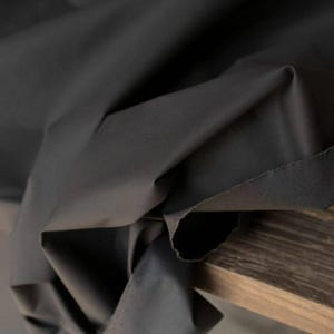 May include: Close-up of a dark gray fabric with visible folds and a raw edge. The fabric appears to be a solid color, possibly cotton or a similar material. The fabric is draped over a wooden surface.
