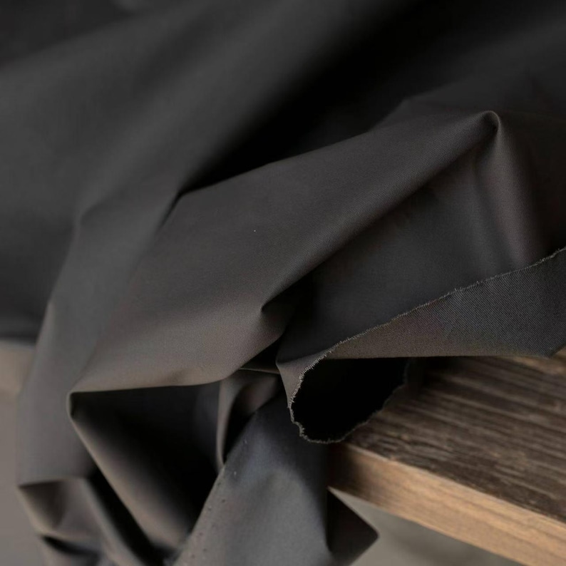 May include: Close-up of a dark gray fabric with visible folds and a raw edge. The fabric appears to be a solid color, possibly cotton or a similar material. The fabric is draped over a wooden surface.