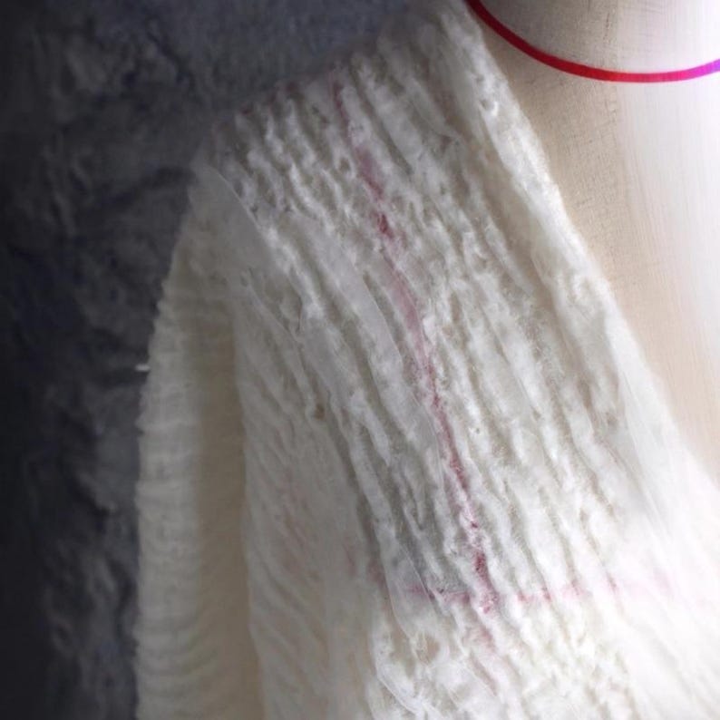 Op de afbeelding: Close-up van een witte, getextureerde stof met verticale ribbels en subtiele roze accenten. De stof lijkt gedrapeerd te zijn, met een zachte, vloeiende kwaliteit. De algehele esthetiek is delicaat en luchtig.