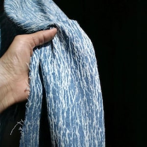 May include: Close-up of a blue and white textured fabric. The fabric has a woven pattern with vertical white lines against a blue background. The fabric is being held, with the edges visible.
