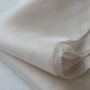 Può includere: Primo piano di un tessuto piegato, bianco sporco. Il materiale sembra essere trasparente e leggero, con pieghe visibili e sottili variazioni di tono. Il tessuto ha un aspetto morbido e fluido, suggerendo un tessuto delicato.