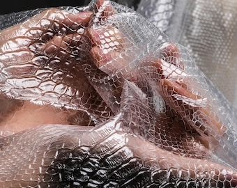 Puede incluir: Primer plano de un material translúcido y texturizado con un patrón de piel de serpiente. El material está drapeado y doblado, creando sombras y reflejos. El efecto general es una apariencia brillante e iridiscente.