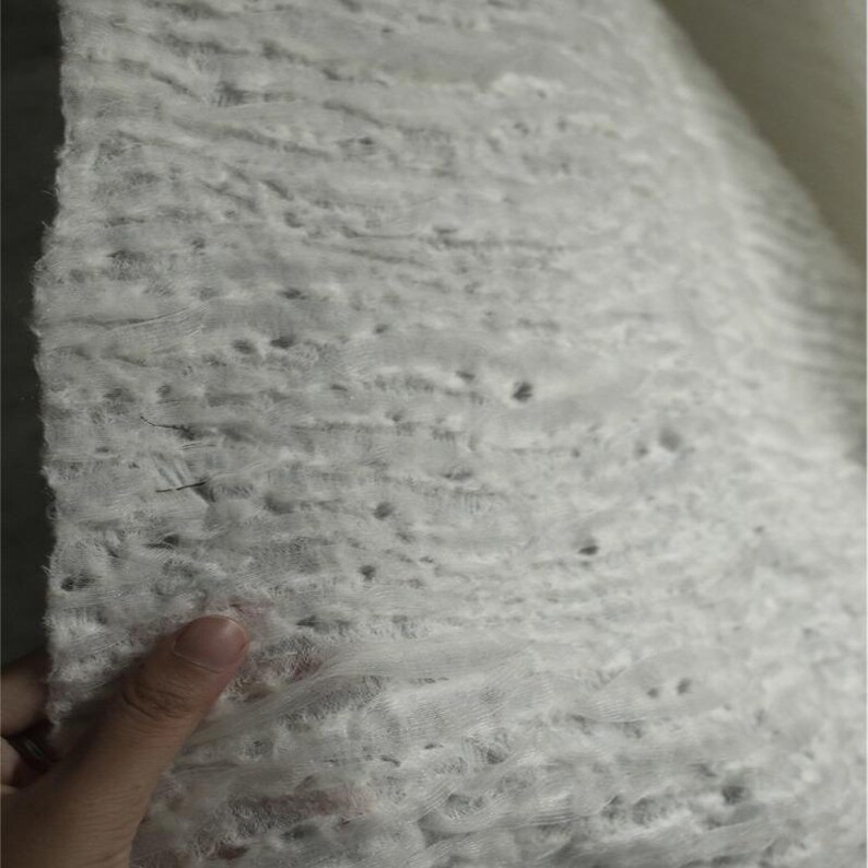 Op de afbeelding: Close-up van een getextureerde, gebroken witte stof. Het materiaal heeft een gelaagde uitstraling met zichtbare gaten en een enigszins ongelijkmatig oppervlak. De stof wordt vastgehouden, waardoor de delicate textuur en het potentieel voor diverse knutselprojecten zichtbaar worden.