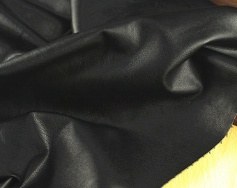 May include: Close-up of a piece of black leather fabric. The leather has a textured surface with subtle variations in tone, and is slightly creased, suggesting flexibility. The lighting highlights the natural grain and sheen of the material, giving it a rich appearance.