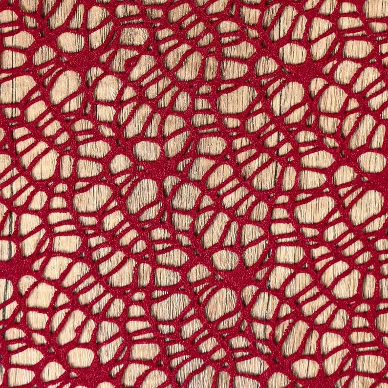 May include: Close-up of a red, open-weave fabric with a pattern of interconnected, irregular shapes. The fabric is set against a light-colored, wood-grain background, creating a contrast in textures and colors. The design resembles a network or web.