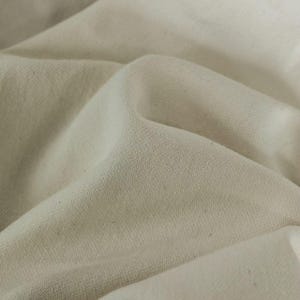 May include: Close-up of a cream-colored fabric with a textured surface. The fabric is folded, creating soft shadows and highlights. The material appears to be a medium-weight textile, possibly cotton or a similar natural fiber.