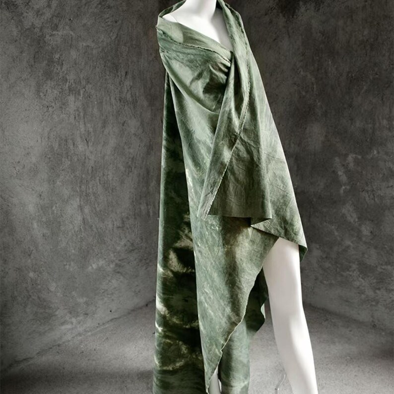 Puede incluir: Una prenda verde oliva drapeada sobre un maniqu&iacute;. La tela tiene un efecto marmoleado, te&ntilde;ido anudado con acentos verdes y blancos m&aacute;s claros. La prenda es fluida y parece ser un chal o envoltura, con una apariencia texturizada.