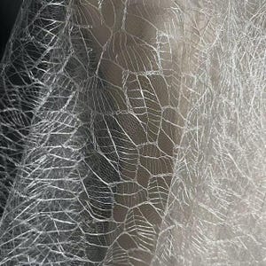 May include: Close-up of a silver fabric with a textured, web-like pattern. The material has a reflective quality, with light and dark areas creating depth. The design resembles interconnected veins or a network.