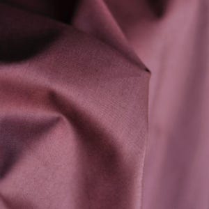 May include: Close-up of a fabric with a deep mauve color. The fabric is folded, creating shadows and highlights that emphasize its texture. The material appears to be smooth and possibly satin or silk.
