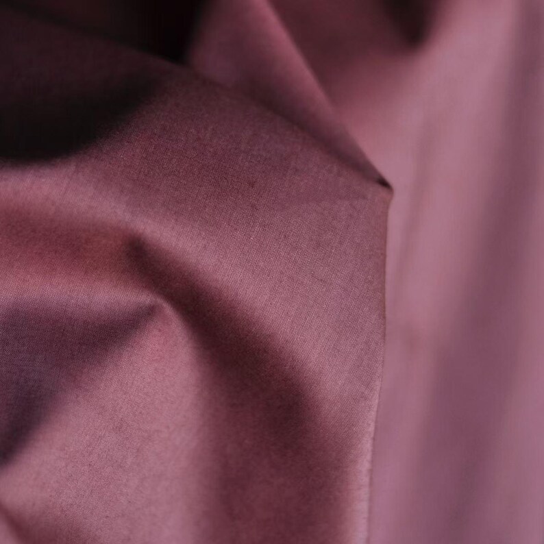 May include: Close-up of a fabric with a deep mauve color. The fabric is folded, creating shadows and highlights that emphasize its texture. The material appears to be smooth and possibly satin or silk.