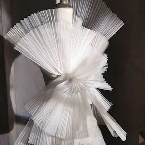 Könnte beinhalten: Ein weißes, mehrschichtiges Kleidungsstück mit aufwendigen Falten. Das Design zeichnet sich durch ein strukturiertes Oberteil und kaskadierende Rüschen aus, die einen voluminösen und strukturierten Effekt erzeugen. Das Kleid wird auf einer Schaufensterpuppe präsentiert, die seine elegante Silhouette zeigt.