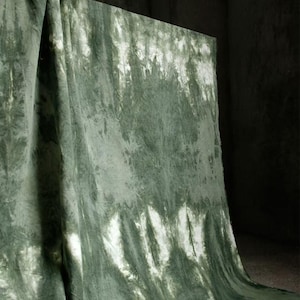 Puede incluir: Una tela drapeada con un patr&oacute;n tie-dye en tonos verdes y blancos. La tela est&aacute; doblada y cuelga en curvas suaves y fluidas, creando una apariencia texturizada. La paleta de colores es apagada, con una mezcla de verdes claros y oscuros.
