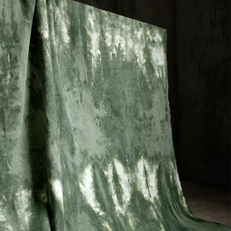 Puede incluir: Una tela drapeada con un patr&oacute;n tie-dye en tonos verdes y blancos. La tela est&aacute; doblada y cuelga en curvas suaves y fluidas, creando una apariencia texturizada. La paleta de colores es apagada, con una mezcla de verdes claros y oscuros.