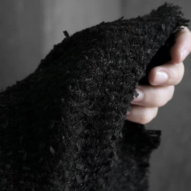 May include: Close-up of a black, textured fabric with a frayed edge. The fabric appears to be thick and possibly knitted or woven, with a rough, uneven surface. The image is focused on the texture and detail of the material.