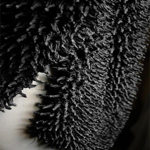 May include: Close-up of a black, textured textile. The fabric has a dense, looped pile, creating a fuzzy, three-dimensional effect. The dark color and tactile surface suggest a focus on texture and material. The textile appears to be a scarf or a similar garment.