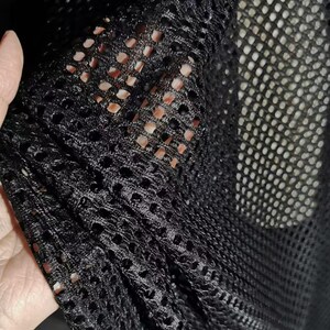 Könnte beinhalten: Nahaufnahme von schwarzem Mesh-Gewebe mit einem gitterartigen Muster. Der Stoff wird hochgehalten und gibt den Blick auf die darunterliegende Haut frei. Das Mesh hat kleine, gleichmäßig verteilte Löcher, die einen durchsichtigen Effekt erzeugen.