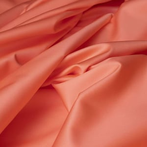 May include: A close-up shot of coral-coloured fabric with gentle folds and a glossy sheen. The fabric appears smooth and flowing, with highlights and shadows adding depth. The material is likely suitable for clothing or home decor.