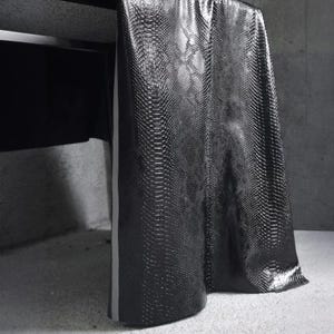 Może przedstawiać: Czarna, teksturowana tkanina z wzorem sk&oacute;ry węża zwisa z ciemnej powierzchni. Materiał jest udrapowany w miękkie fałdy, odbijając światło. Cienki, jasny pasek biegnie pionowo wzdłuż krawędzi.