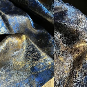 Könnte beinhalten: Nahaufnahme eines schimmernden Stoffes mit strukturierter Oberfläche. Der Stoff weist eine Mischung aus dunkelblauen, goldenen und silbernen Farben auf, die ein luxuriöses und elegantes Aussehen erzeugt. Das Material hat einen subtilen Glanz, der Tiefe und visuelles Interesse verleiht.