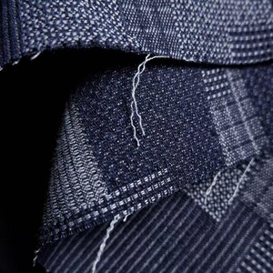 May include: A dark blue fabric with a woven texture featuring a grid-like pattern of lighter blue and white threads. The material shows a folded edge with visible loose white fibers along the seam, highlighting the dense, interlaced weave structure.