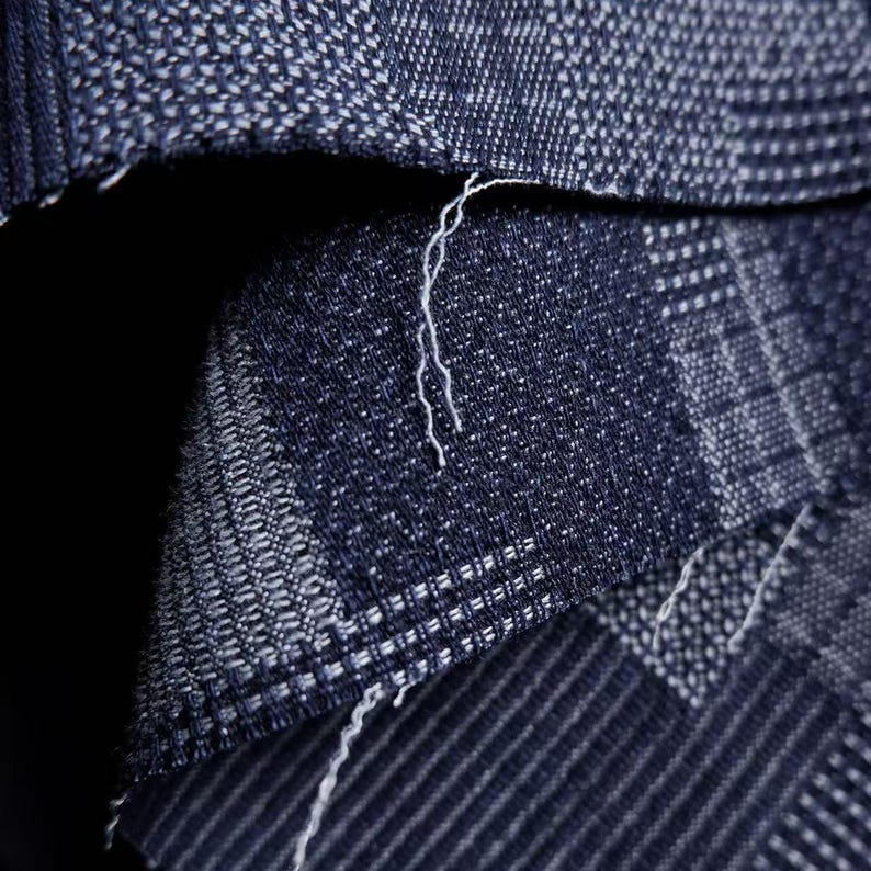 May include: A dark blue fabric with a woven texture featuring a grid-like pattern of lighter blue and white threads. The material shows a folded edge with visible loose white fibers along the seam, highlighting the dense, interlaced weave structure.