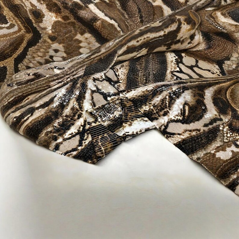 May include: Close-up of a fabric with a snakeskin pattern in shades of brown, tan, and black. The fabric has a textured appearance and is embellished with silver sequins along the edges, adding a touch of sparkle to the design.